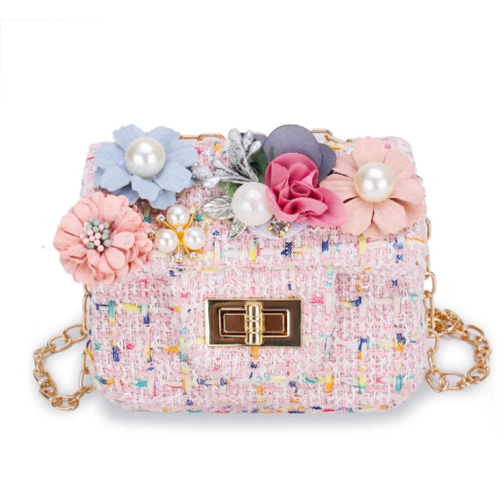 A small pink tweed shoulder bag with colorful embroidered flecks, decorated with large fabric flowers, faux pearls, and a gold-tone clasp and chain strap, shown on a plain white background.